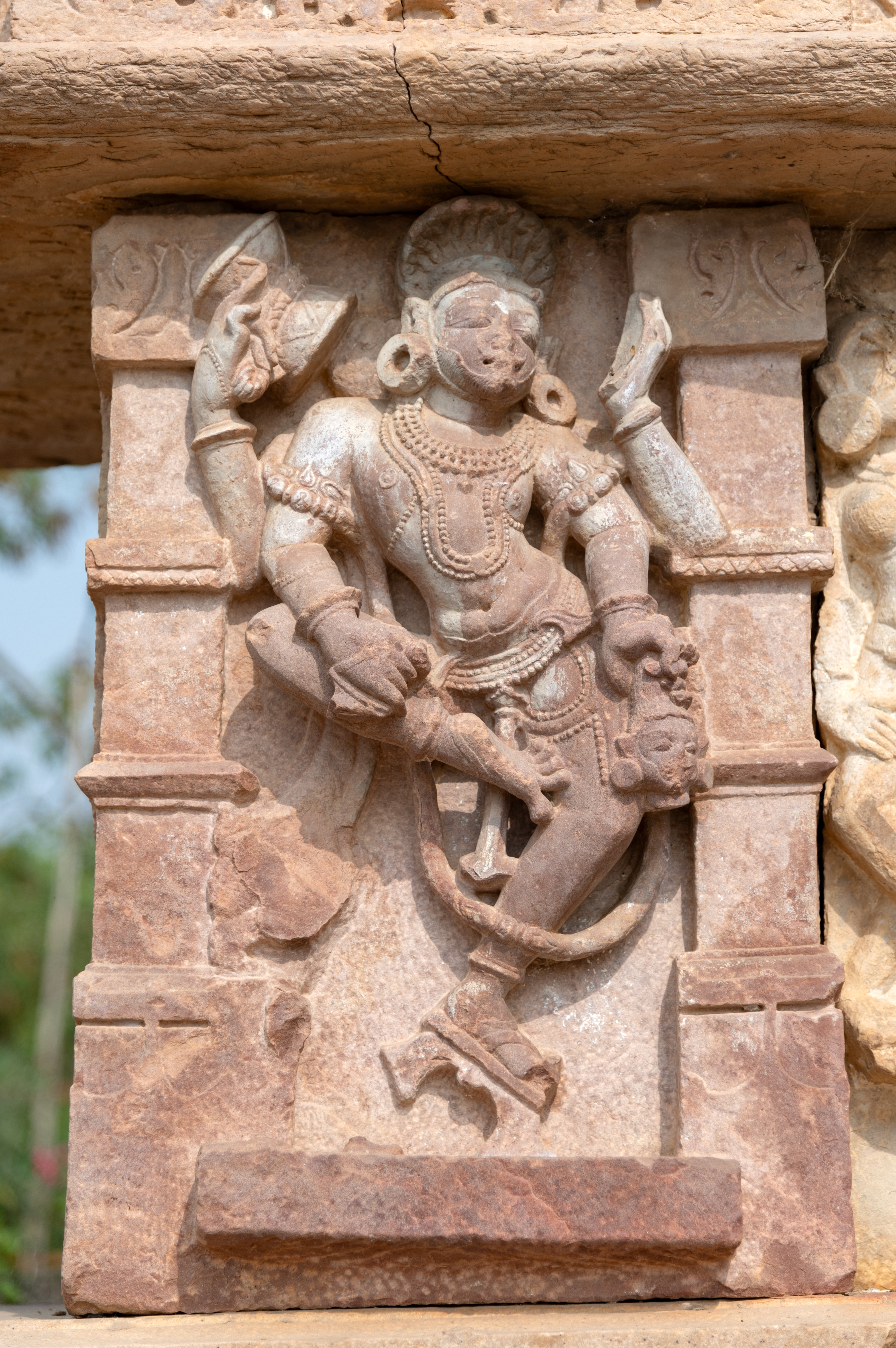 Bhairava is portrayed dancing, his four arms moving in a divine rhythm. Notably, Bhairava wears unique footwear with a distinct heel. His left foot, adorned with a sandal, rests firmly on the ground, while his right foot is raised inward, adding dynamism to his celestial dance. In his upper hands, Bhairava holds a damru (drum) and a bowl. His lower left hand holds a severed human head, while his lower right hand grasps an object resembling a musala (pestle). His appearance is marked by a highly raised hairstyle, a moustache and a flowing beard.