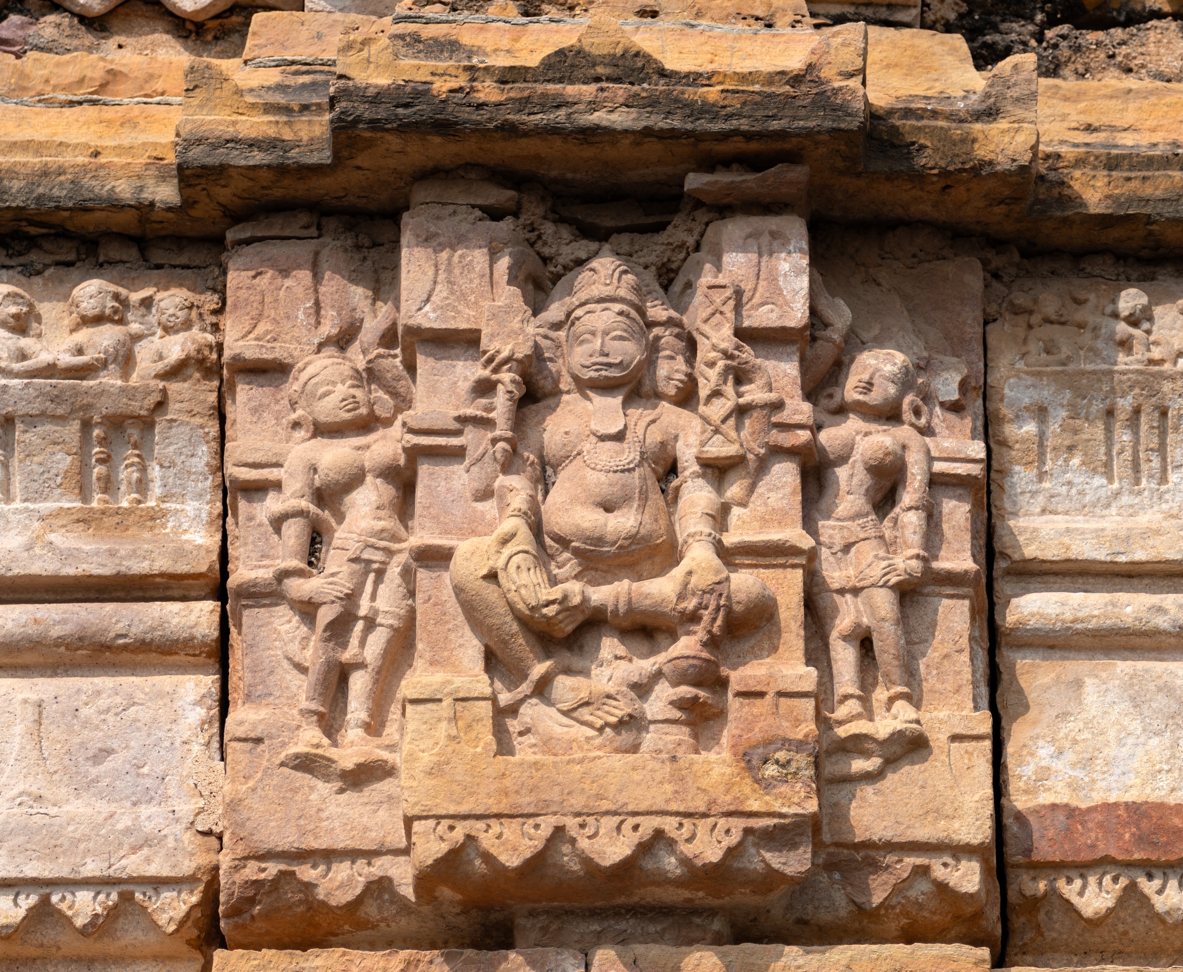 Image 26: On one facet of the shikhara (superstructure), Brahma sits in divine contemplation. Portrayed with three faces and four arms, Brahma holds a shruka (sacrificial ladle) in his upper left hand, while his upper left hand holds a pustaka (sacred text). His lower right hand is in the varada hasta gesture, while his lower left hand clasps a kamandalu (water pot), representing purity and sustenance. He is adorned with a jatamukuta (crown of matted hair) and jewellery. His long eyes, straight nose, and full lips convey a sense of timeless grace, while his long beard adds to his aura. Bare-chested, Brahma wears a broad yajnopavita (sacred thread), symbolizing his connection to spiritual and ritualistic wisdom.