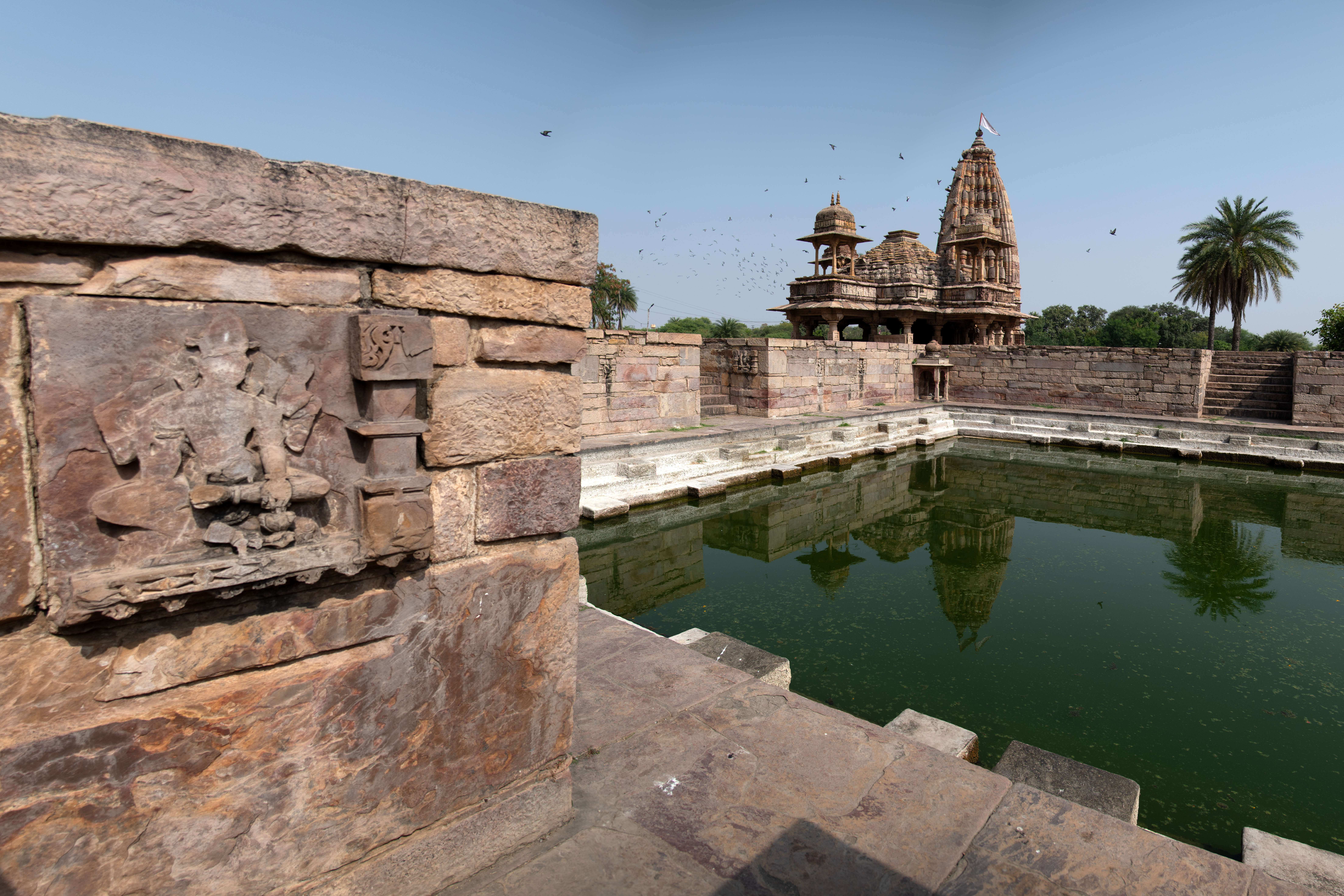 Image 2: The Mandakini Kund reflects the vast skies, with birds in flight, and the temple spires creating a peaceful scene. One can see the depiction of a deity on the wall, which may have been a later addition.