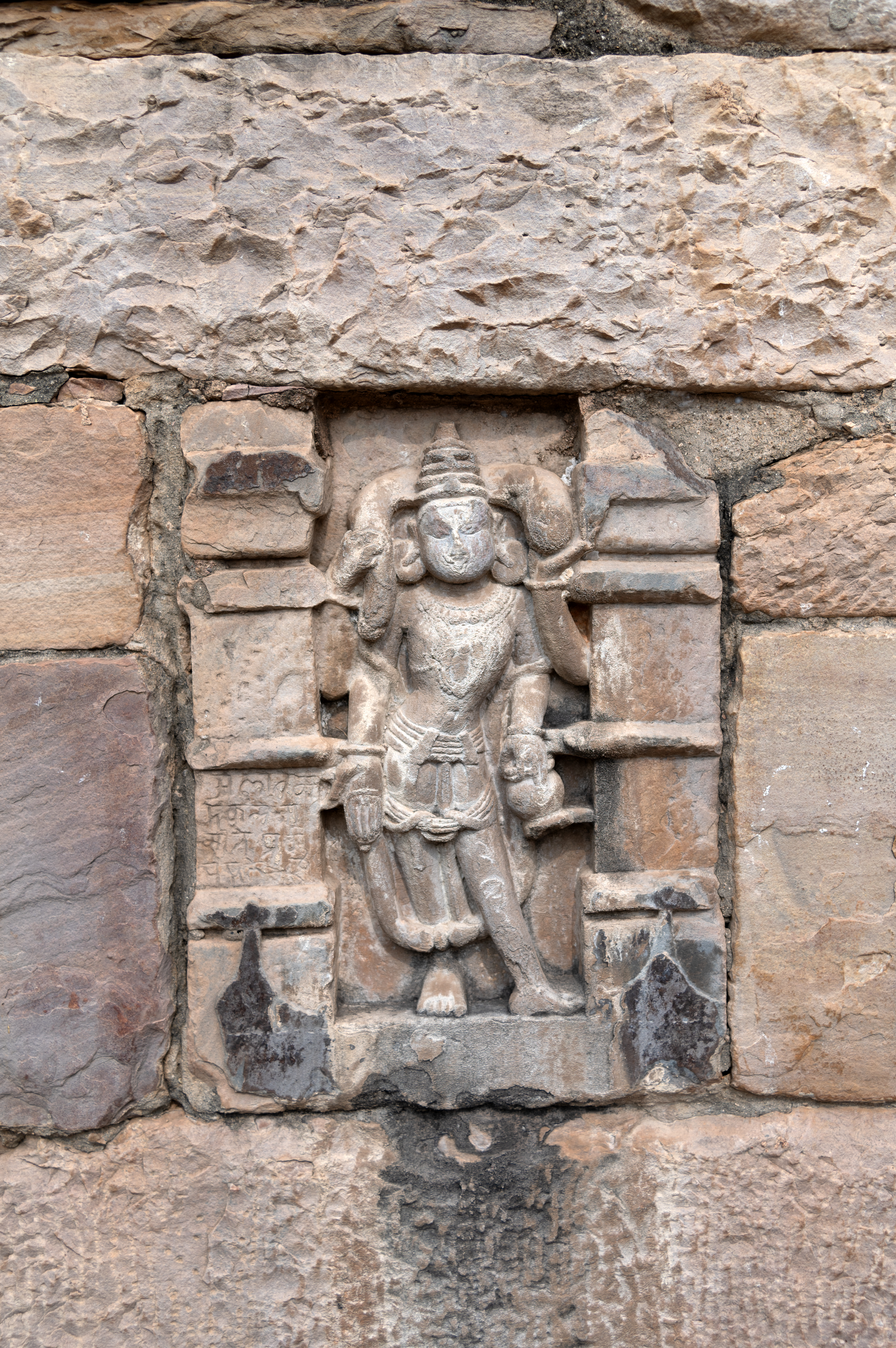 Image 15: In addition to the main deities and pantheon, several sculptures of dikpalas (guardians of directions) are also present. One of these dikpalas is Kubera, with two sculptures visible on the enclosure wall. Both depictions show Kubera crowned, with four arms and standing. He holds a money bag in his upper arms, while his lower right hand is in varada hasta (boon-giving gesture) in both sculptures. In his other lower arm, he carries a kamandalu (water pot) in one sculpture and a bowl-like container in another. The only distinction between the two sculptures is that one portrays a mature male deity with a beard and moustache, while the other depicts a youthful deity with a slim, toned body.