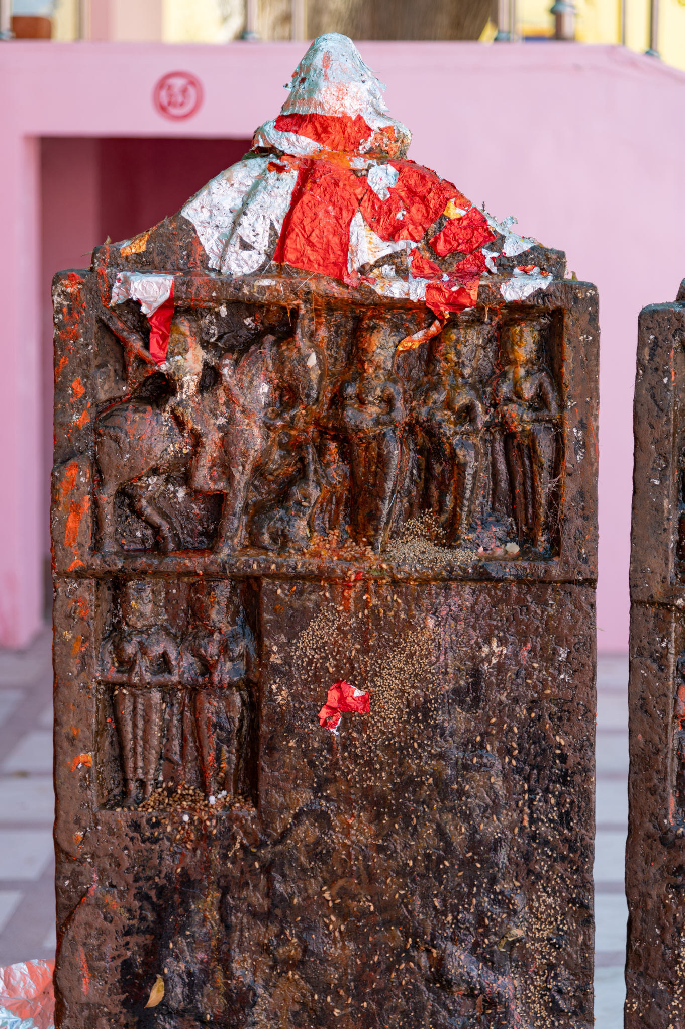 The topmost portion of the hero stone slab has a depiction of four figures, three standing on foot and one mounted on a horse. This is a depiction of the ‘hero’ who embarked on an expedition, fought against enemies, and ultimately lost his life on the battleground. The erection of the hero stones is a tribute to honour his heroic deeds and sacrifice. He is also accompanied by some other figures, but the vermilion and oil make them difficult to identify. The three figures standing in front of the hero may represent celestial damsels who are shown bowing down to him. However, due to the presence of vermilion and oil on the stone, discerning their identities is challenging.