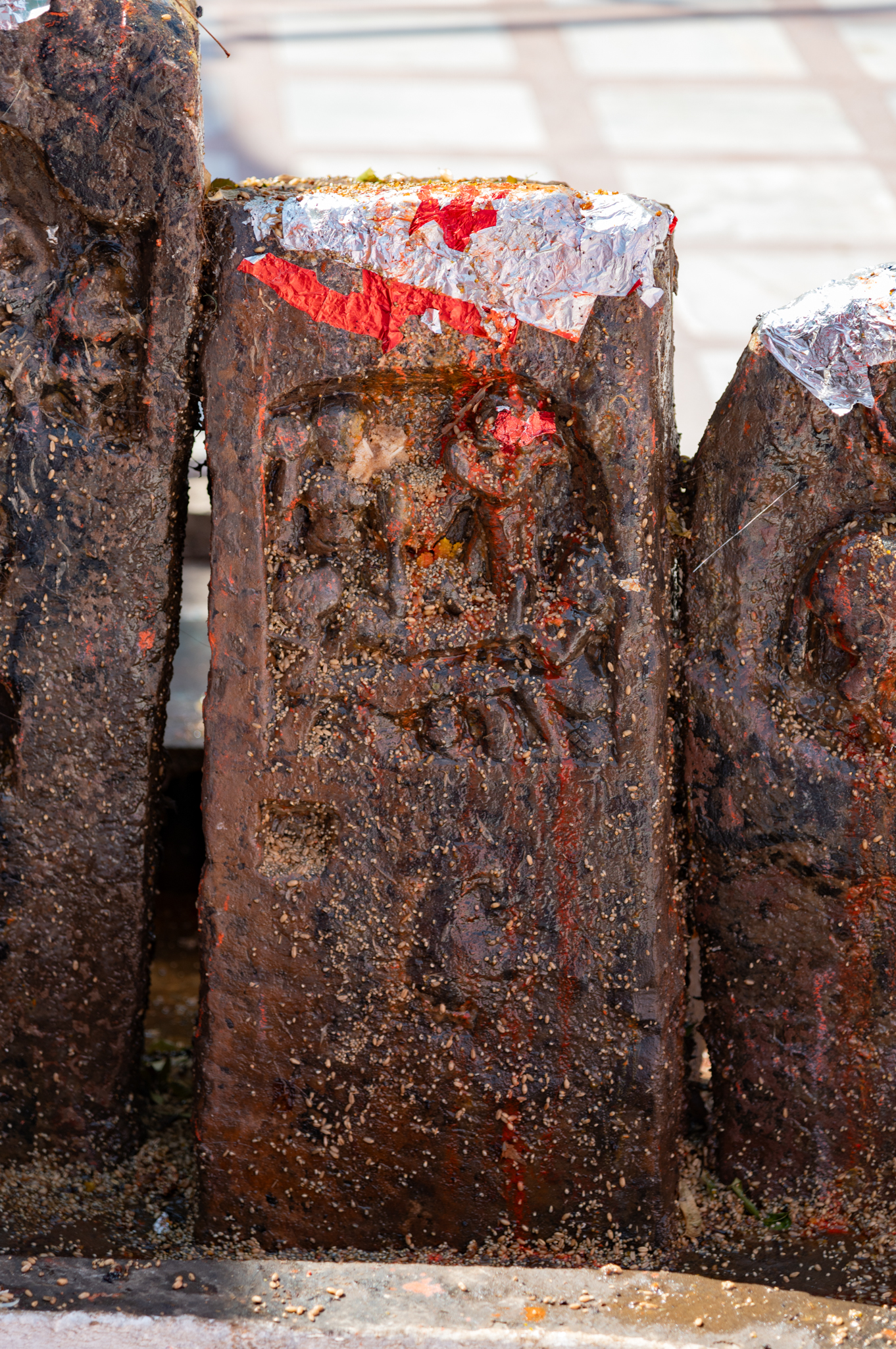 Seen here is a small-sized hero stone, similar in dimensions to the adjacent hero stones. It is a rectangular stone slab without a triangular finial. The panel depicting the hero and his journey to the heavenly abode differs slightly from others. Here, there is no figure mounted on horseback; instead, there is a supine human figure next to another figure standing with folded hands. The depiction likely indicates the hero’s death and his attainment of moksha (freedom from the eternal cycle of life, death, and rebirth). This unique portrayal is not found on any other hero stone fragments in the Suswani Mata Temple.