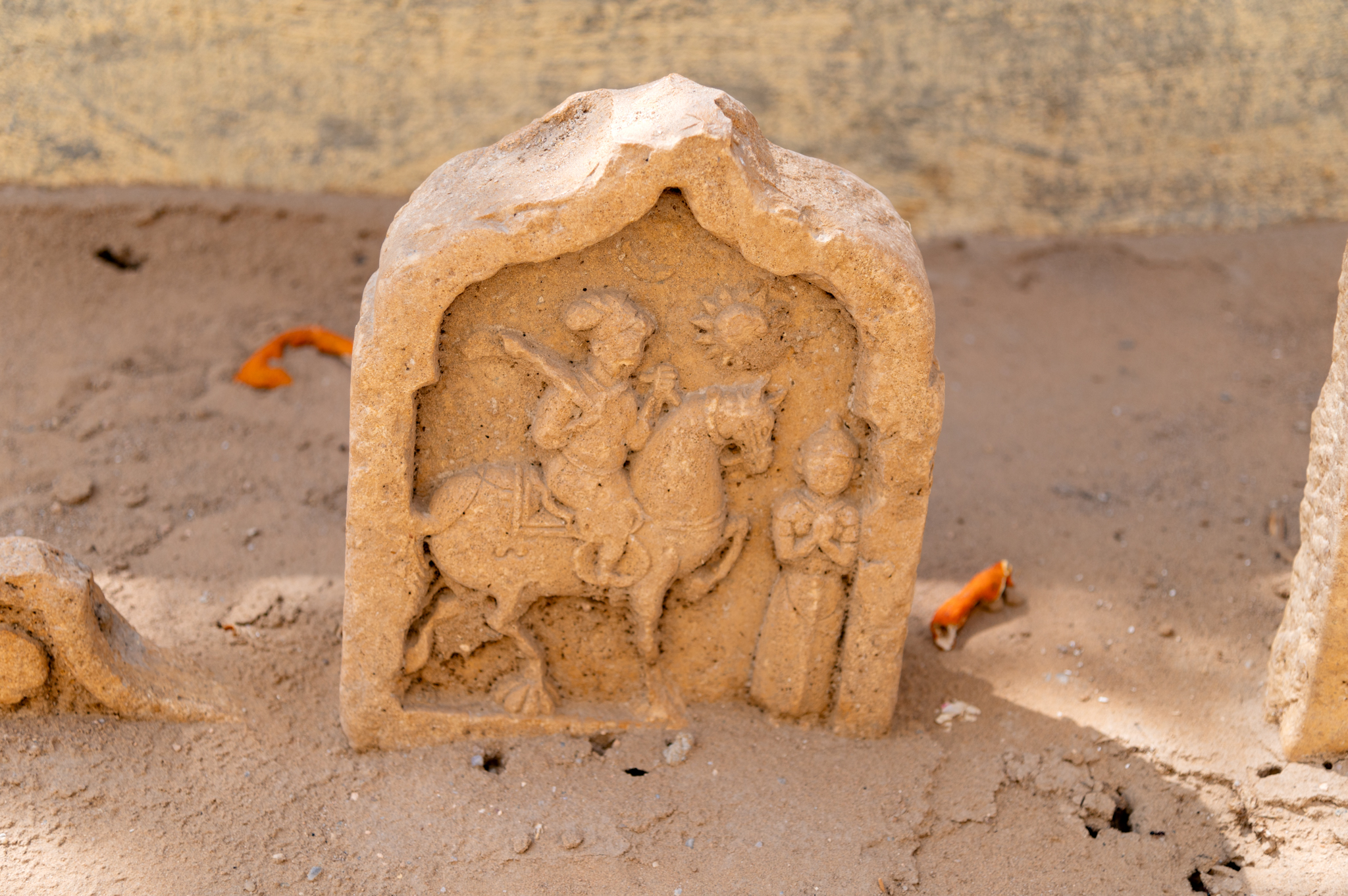 Seen here is an example of a hero stone without an inscription, showcasing a slightly different style. The niche inside which the hero is depicted is a low-relief niche with an arch design. The drapery of the hero and the celestial damsel accompanying him is uniquely rendered. The hero is depicted wearing a distinctive headgear resembling those worn in the Rajasthan region. In the corner of the niche, there is a sun motif, possibly symbolizing that the hero’s valour will endure eternally.