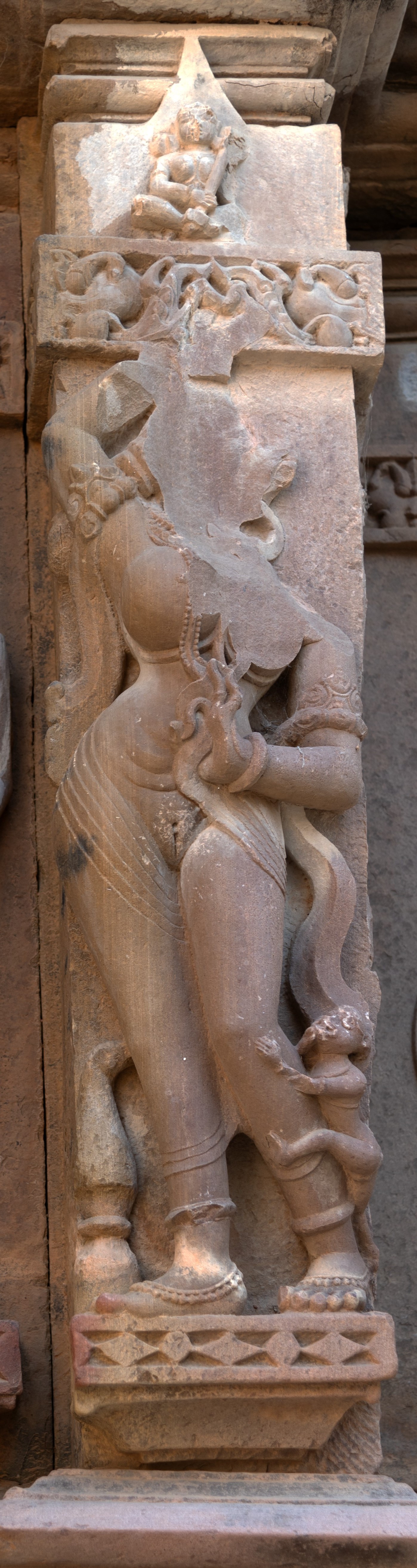 The surasundari (celestial damsel), presently faceless, stands gracefully with her voluptuous body adorned with exquisite ornaments on the eastern wall of the garbhagriha (sanctum sanctorum). Although her right hand is raised and damaged, she holds a fine scarf in her left hand near her belly, wrapping it around her arms. Her lower body is veiled by a fine translucent garment. A small creature is depicted moving upward on her left leg. The surasundari vividly embodies sensual beauty, in stark contrast to the tranquil and composed posture of the ascetic. There is a kalasha (water pot) placed at the bottom near her feet with foliage, symbolizing abundance and growth, as does the female body.