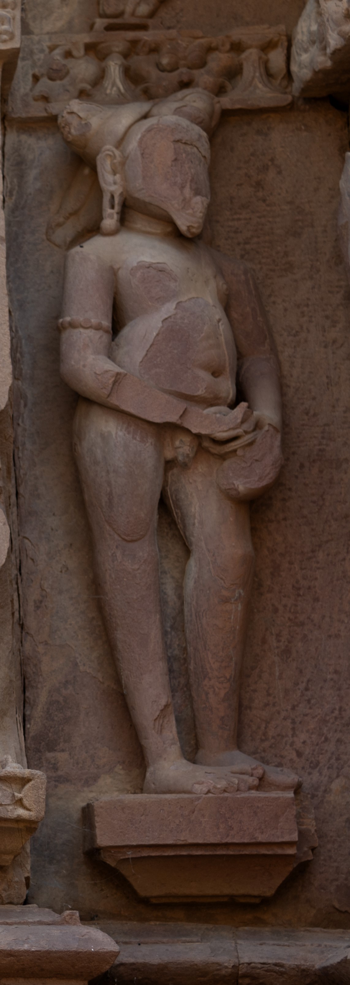 In an alternative portrayal of the ascetic, he is depicted standing, facing slightly to his left. He has a jatabhara (matted hairdo) atop his head and a lengthy moustache. The features of his face have eroded, but two notable ones are his elongated earlobes and the rudraksha (dried stones or seeds of the Elaeocarpus ganitrus associated with Shiva iconography) armband. One of his hands clasps a pot while his fingers are delicately positioned atop it. Notably, his phallus is exposed in this depiction.