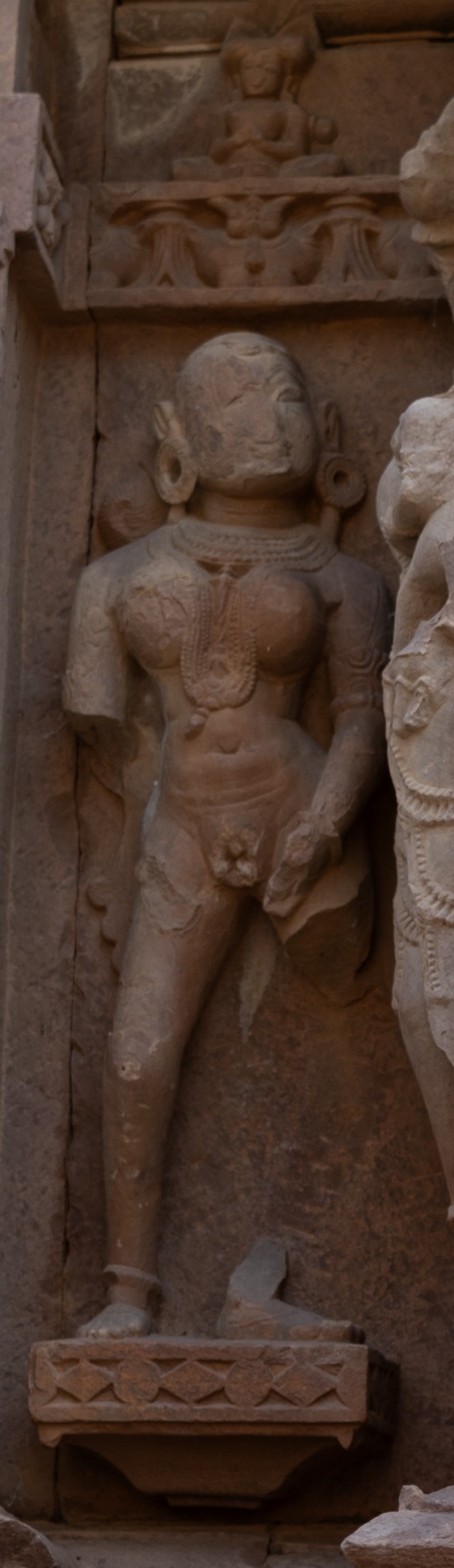 On the northern wall, there is a depiction of a surasundari (celestial damsel) with an elaborate hairdo fashioned into a bun. She stands tall and slender, bedecked with substantial jewellery. One of her hands is fractured, while the other rests near her exposed reproductive organ. It is worth noticing that both ascetic and surasundari are shown with their revealed genitalia, both representing different natures and standing for opposite symbolism.