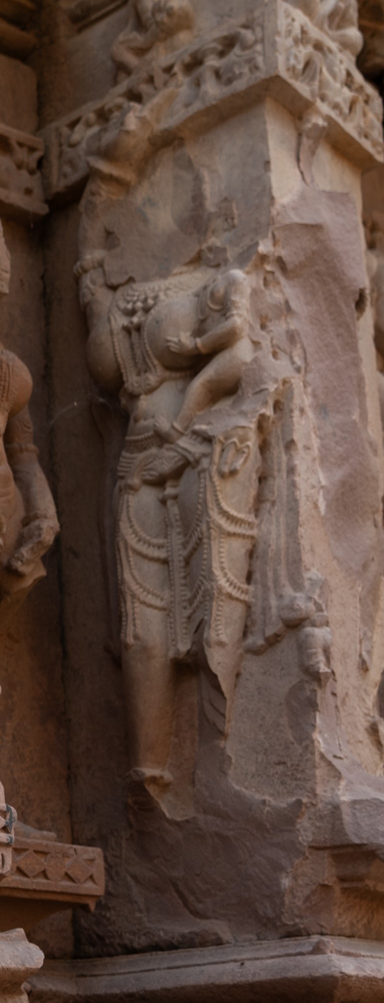 The portrayal of this surasundari (celestial damsel) on the northern wall encapsulates the nurturing essence of femininity. Standing gracefully, she cradles a baby in her arms, although the faces of both mother and child are obscured. The mother has a voluptuous figure adorned with elaborate ornaments. The child is depicted holding her breast, symbolizing the bond of maternal care and sustenance.