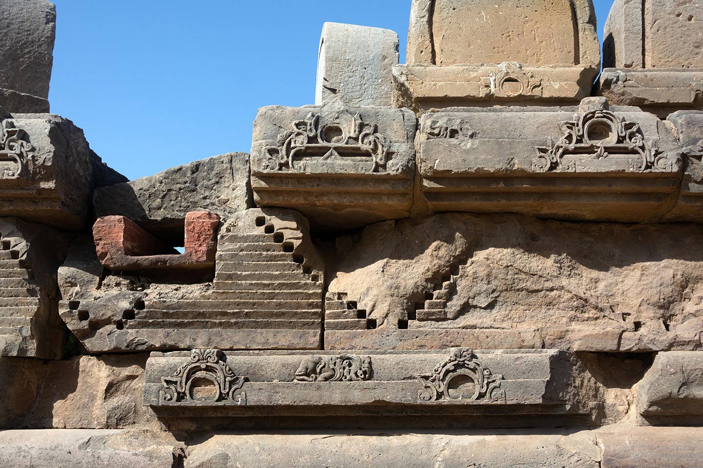 All around the adhisthana are relief carvings of geometric, flora, and fauna motifs. On the left, there is a pranala (water spout) for drainage.