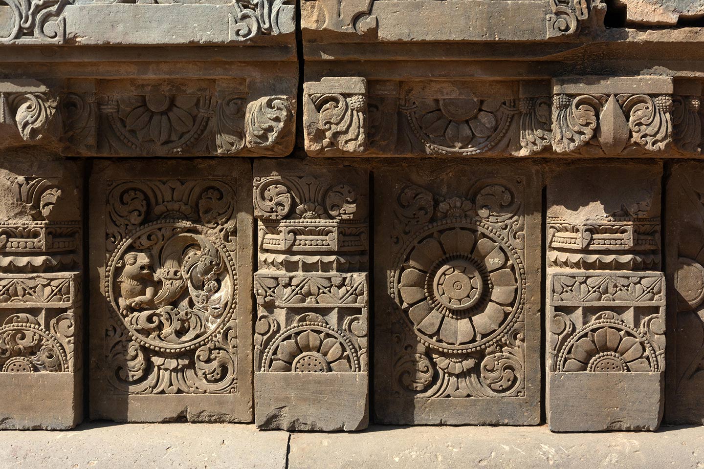 The plaque (on the left) depicts a simha (lion) vyala, complemented by creeper motifs that fill the rest of the medallion. The plaque, on the right, features the ashtadal kamal (lotus with eight petals). Squat pillars, featuring a square base, an octagonal shaft, and a square capital, separate the plaques. The pillars are decorated with ardha padma (half lotus) and kalpa lata (creeper) motifs.