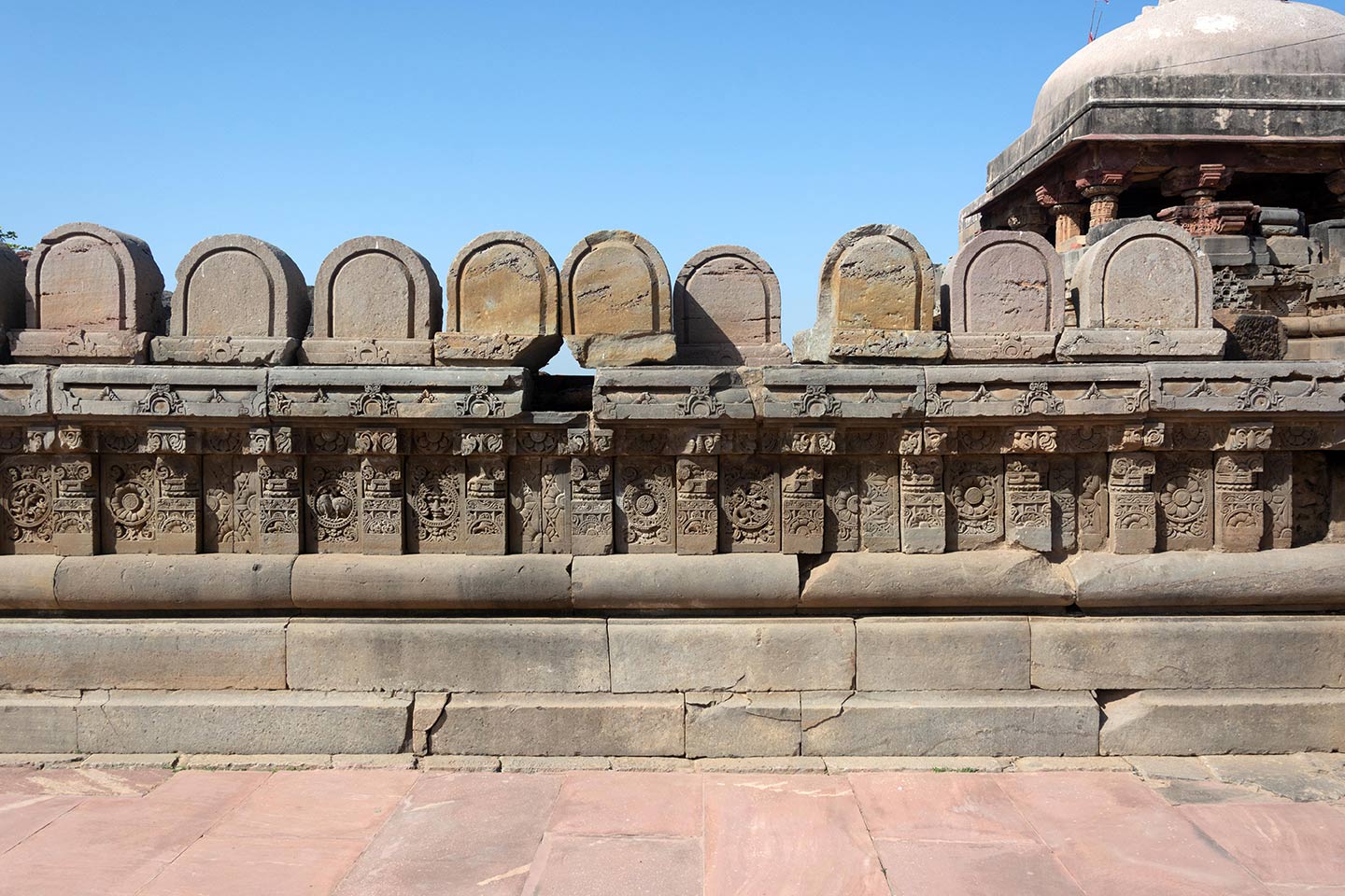 U-shaped rounded stones are assembled on the parapet, but their position or purpose in the original temple is unknown. The central artwork of the plaque is in the form of a circular medallion surrounded by a foliage motif. The plaques are separated by squat pillars which have a square base, an octagonal shaft, and a square capital.
