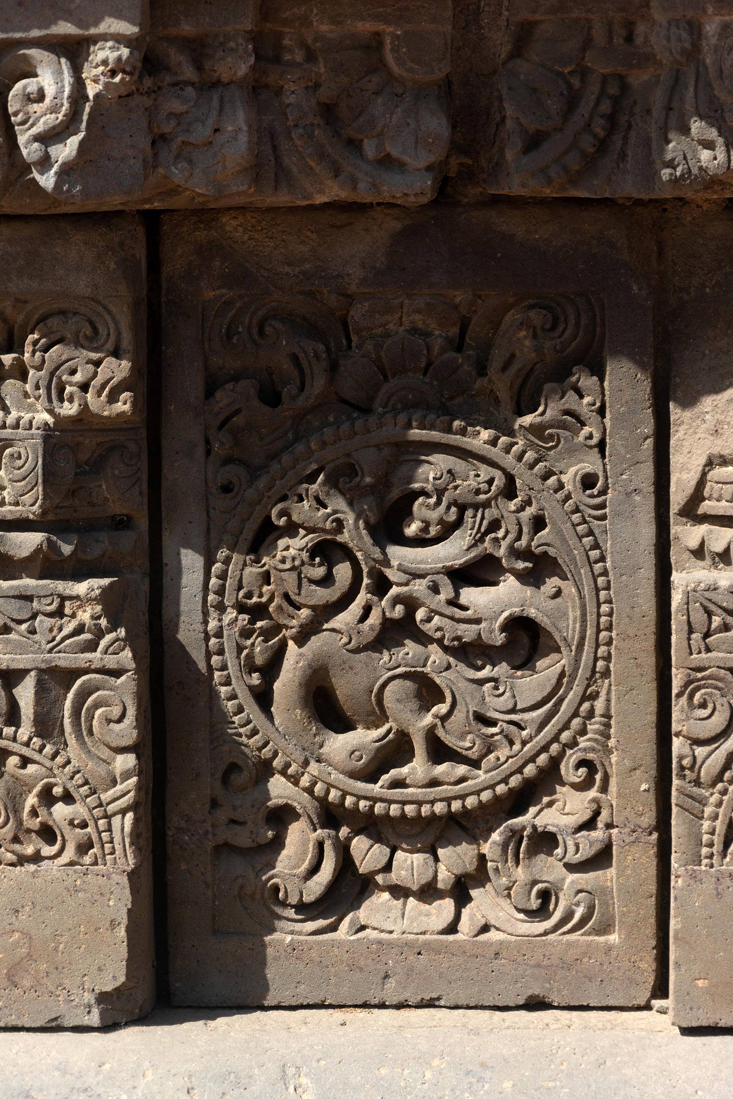 The central artwork of the plaque is in the form of a circular medallion surrounded by a foliage motif. In this medallion, a bird (likely a peacock) with its head bent downwards is integrated with a creeper motif. The rest of the plaque is covered with ardha padma (half lotus) and kalpa lata (creeper) motifs.