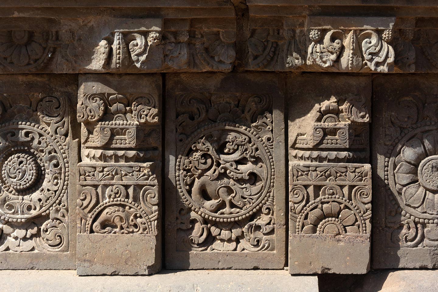 The central artwork of the plaque is in the form of a circular medallion surrounded by a foliage motif. In this medallion, a bird (likely a peacock) with its head bent downwards is integrated with a creeper motif. The plaques are separated by squat pillars which have a square base, an octagonal shaft, and a square capital. The pillars are decorated with ardha padma (half lotus) and kalpa lata (creeper) motifs.