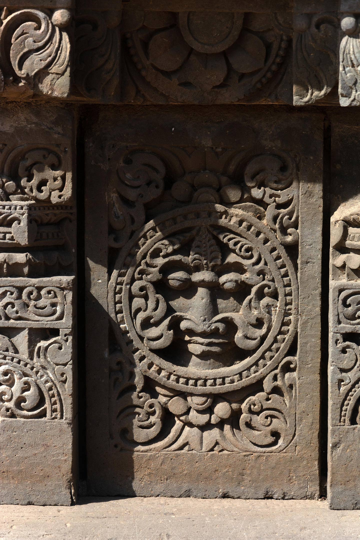 The plaque's central artwork consists of a circular medallion surrounded by a foliage motif. This plaque features a kirtimukha (face of glory) with ardha padma (half lotus) and kalpa lata (creeper) motifs covering the rest of the plaque surface.