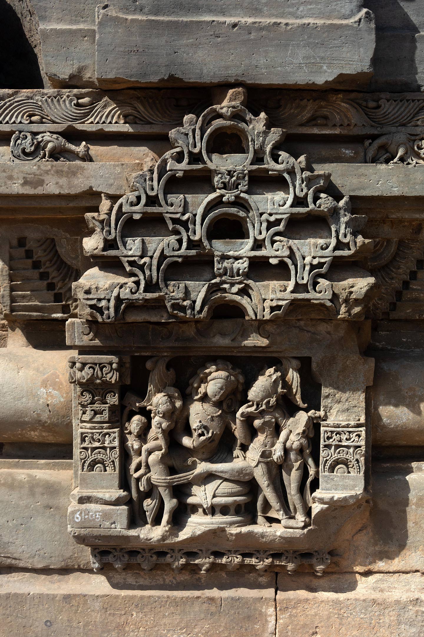 The scene depicts the couple admiring each other, in a moment of shringar rasa (romantic love). The central figure of the man is seated in the lalitasana posture. He is caressing his partner’s chin with his left hand while holding a flower in his right hand, offering it to her. The lady is resting her right hand on the left thigh of the man and her head is tilted upwards, as he looks into her face.