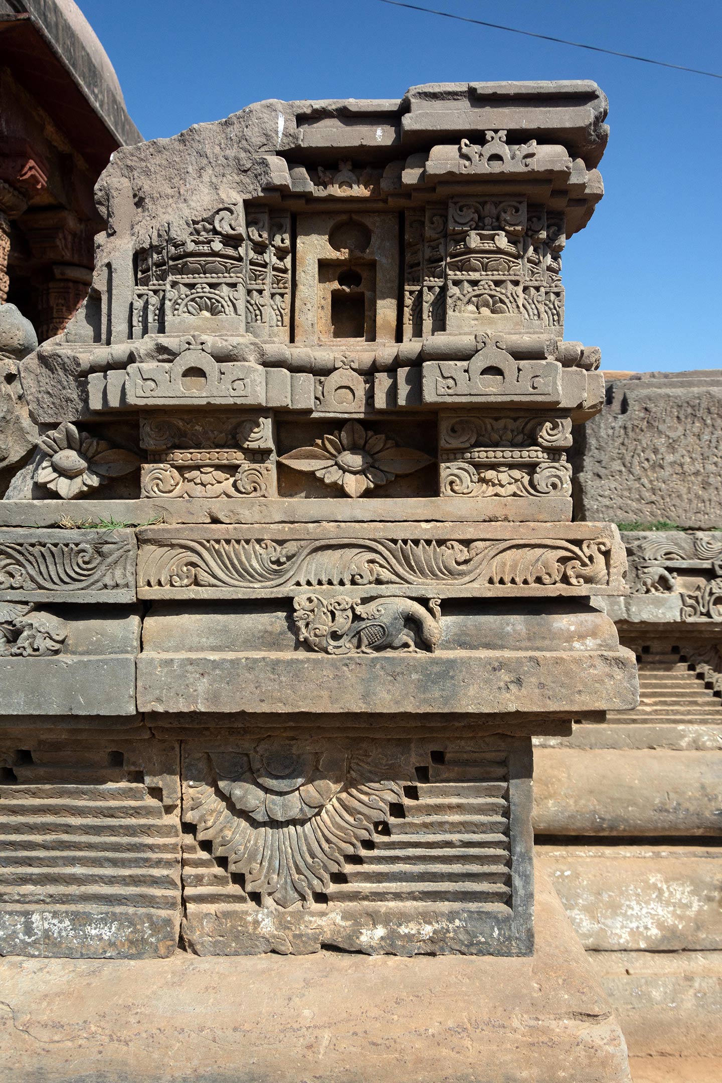 The base has an ardha padma (half lotus) surrounded by geometric patterns. The panel in the middle has an ornate bird, probably a mayura (peacock), bordered by a creeping vine motif. The next two levels are mouldings depicting floral and vine patterns.