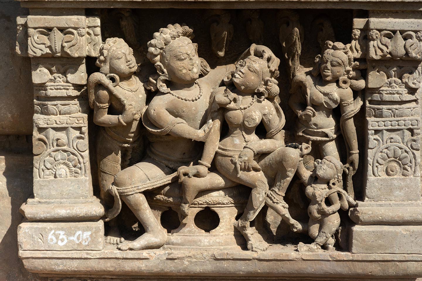 Couples invoking shringara rasa (associated with romance, love, and attractiveness between lovers) are framed in panels with pillars and tiered shikhara with gavaksha (horseshoe) motifs. The central male figure is seated in the lalitasana pose on a raised seat. The central female figure is seated on his left thigh, looking towards the man with her head turned. The man is admiring the beauty of the woman, placing his left hand on her coiffure and inspecting her face with his left hand (missing).