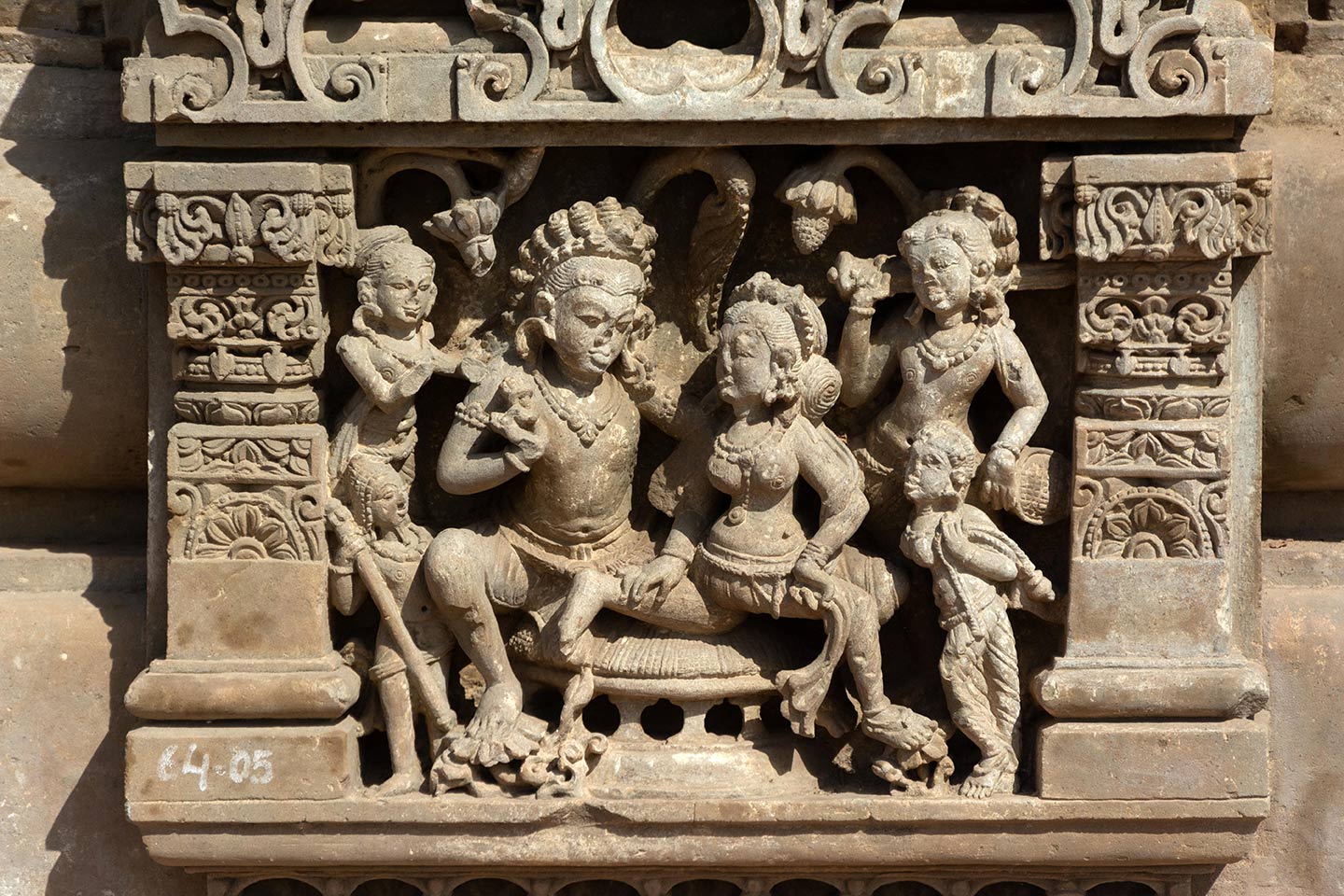 The central female figure is seated on his thigh, looking towards the man with her head turned. Both rest their right feet on blooming lotus flowers. The man is holding a flower in his right hand while the woman gently rests her right hand on his left foot. Trees in the background suggest they are in a grove. The couple is surrounded by four male and female attendants.