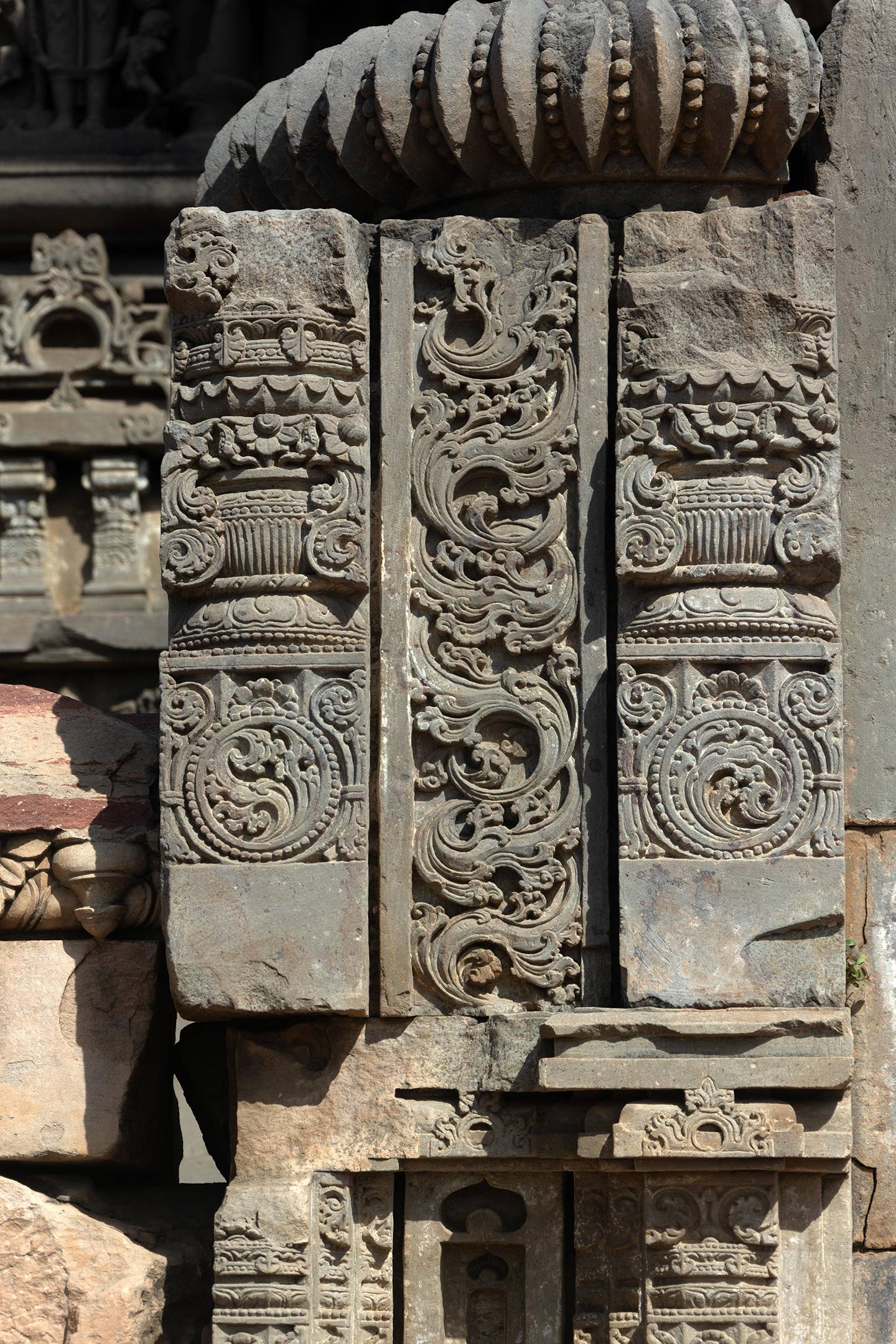 A broken section of the block consists of two pillars that are divided vertically into three sections. The kalpa lata (creeper) motif is at the base, and the purna kalasha (pot of prosperity) is in the center. The top part is damaged, and blooming lotuses separate the three parts. The central part features a detailed carving of a flowing kalpavrisksha (tree of life) motif.