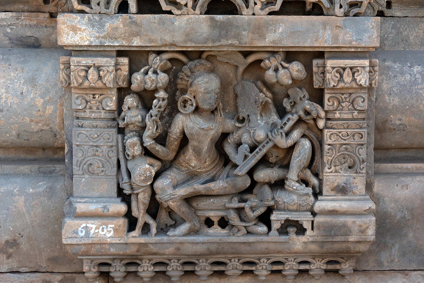 The central male figure and female figures are enjoying a musical concert in a grove. They are seated on circular raised seats, and the male is seated in the lalitasana pose. The female figure is leaning towards the male and playing a stringed musical instrument (likely a veena). They are surrounded by four smaller figures. A male soldier is holding a sword and shield (on the left). One musician is playing the flute (bottom), sitting between the seats.