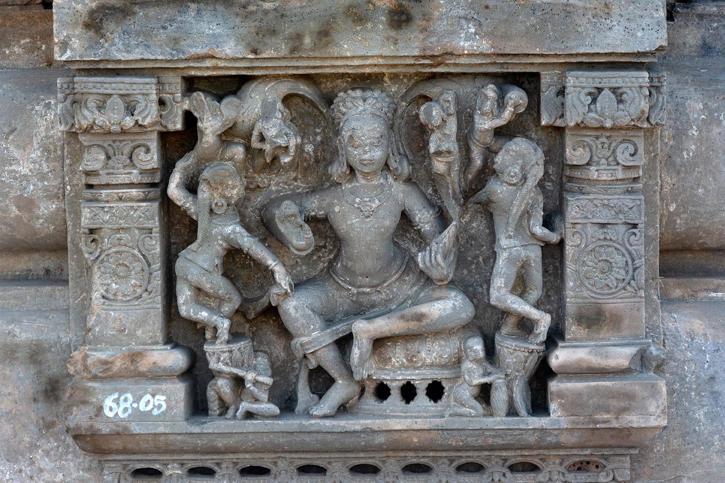 A male figure surrounded by dancers and musicians is framed in panels with pillars and tiered shikhara with gavaksha (horseshoe) motifs. Two female dancers are balancing themselves on big drums on either side of the male figure. Small figures of musicians are holding the base of the drums. The one on the left is playing the flute. Four small figures appear at the top, playing cymbals, two on either side of the male figure.
