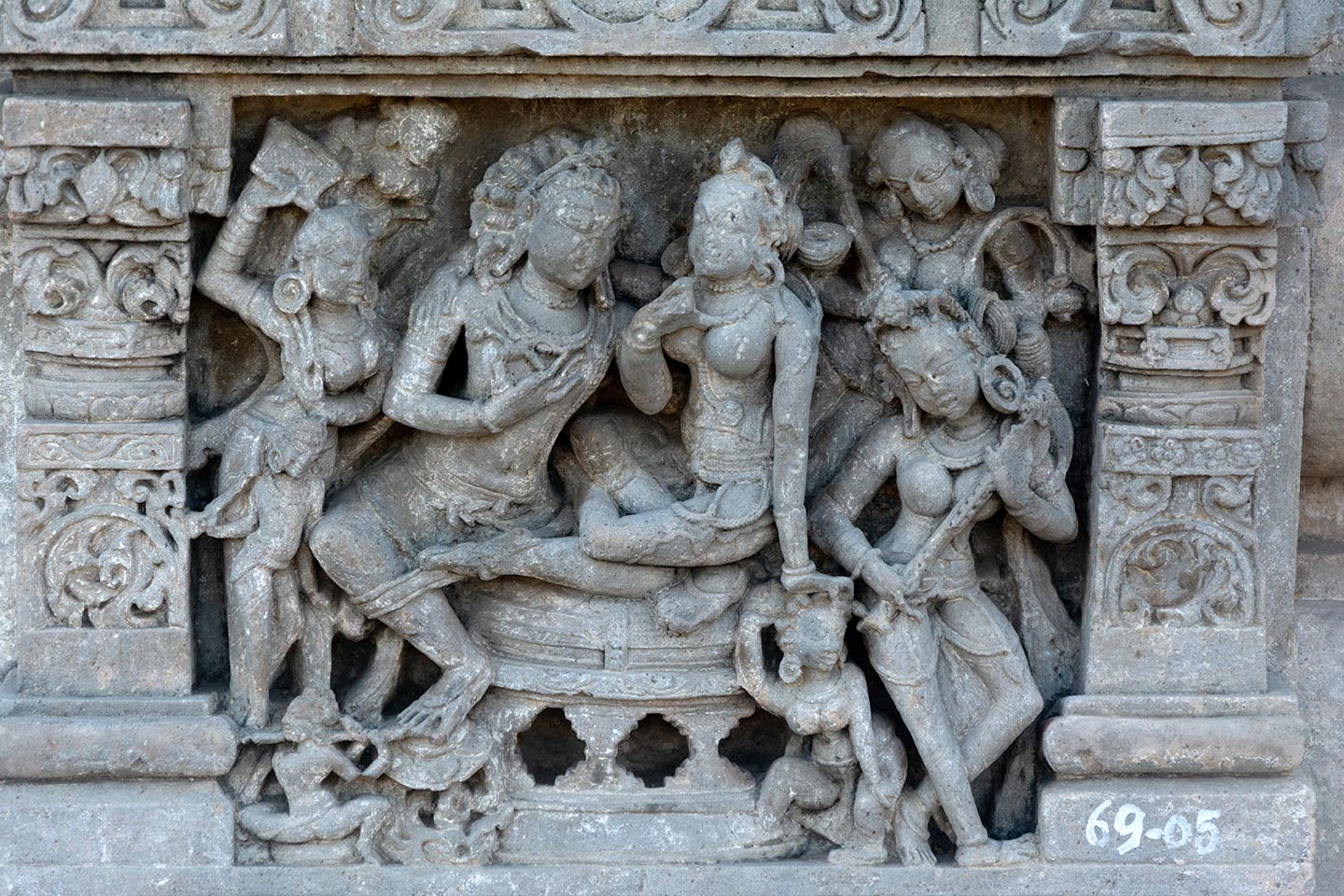 The man is facing the woman and caressing her chin with his left hand and holding a flower in his right hand. The female has her right hand placed over the man’s right shoulder, while her left-hand rests upon the head of a female attendant seated on the ground. The man is resting his right foot on a lotus next to a musician playing the flute (bottom left). Three more attendants appear on the left, with the largest one holding a fan.