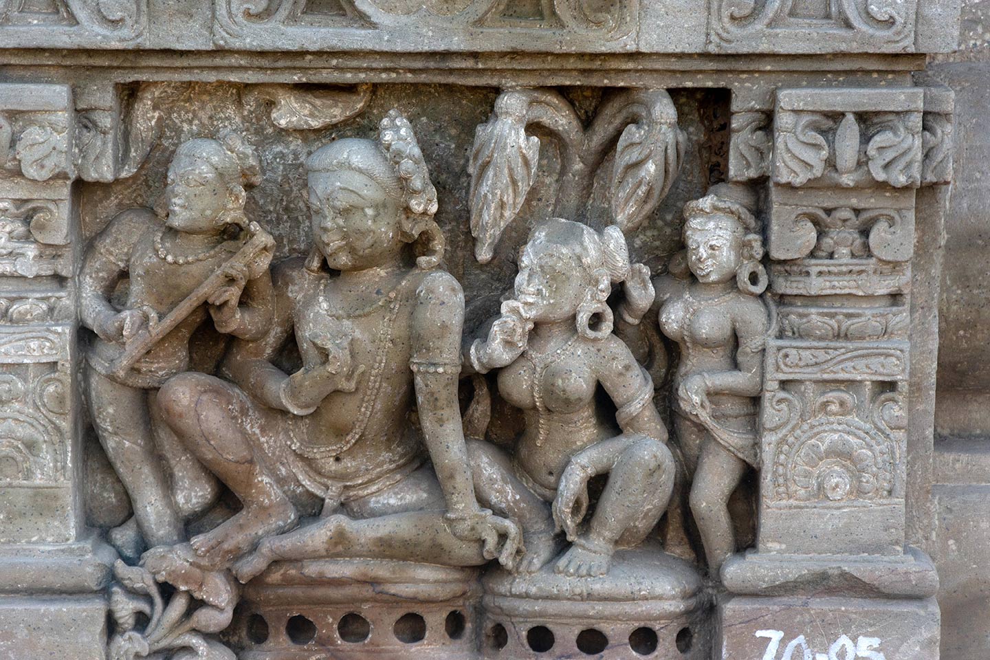 The central male figure is seated in the rajalilasana pose (both legs raised on the seat, palm of the feet together) on a raised seat. The central female figure is seated on the left of the man, on a separate seat, in the malasana (squat) pose. Male and female attendants flank the central figure from the left and right respectively. All the figures are watching a performance (not in the frame) and looking in unison towards the left.
