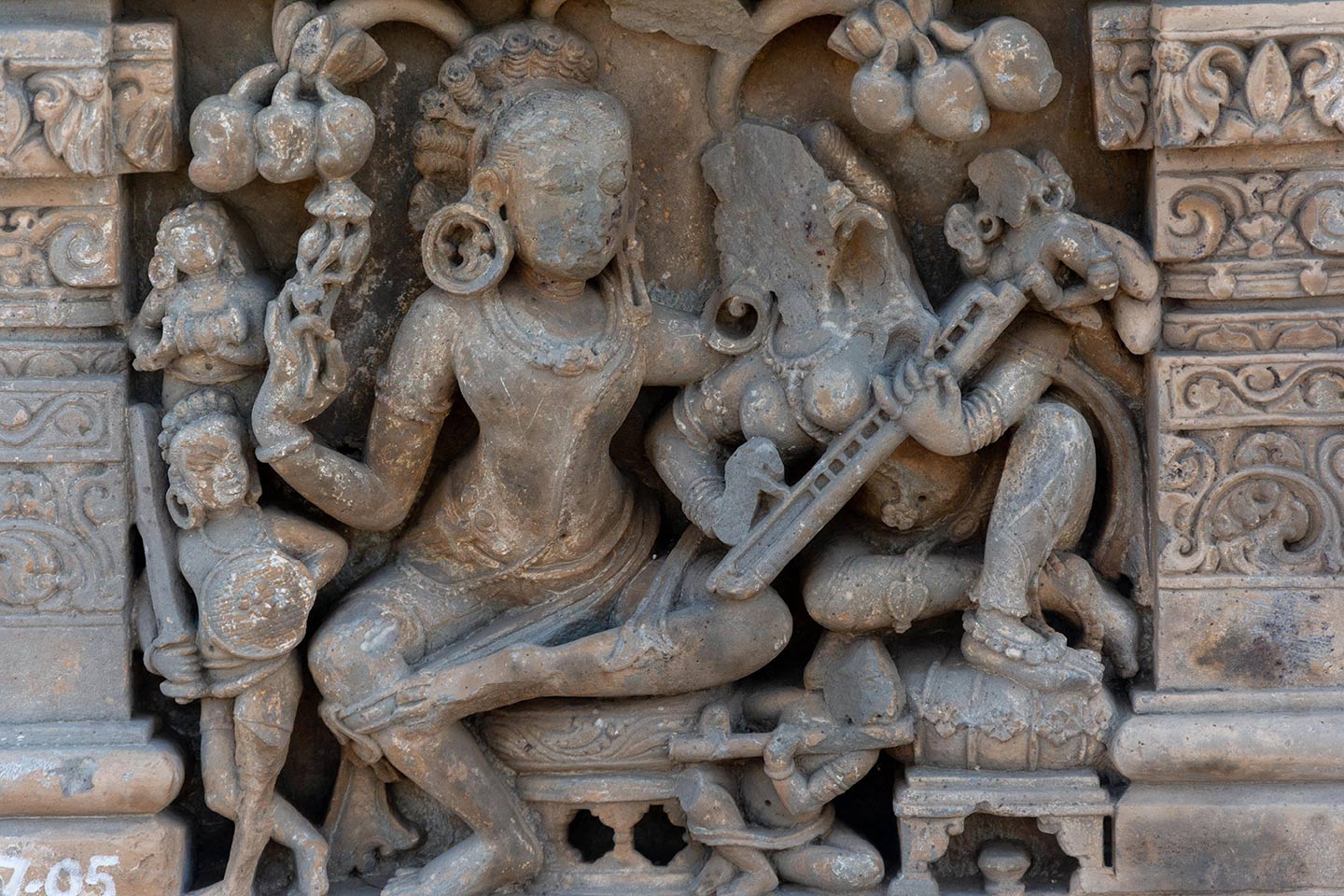 The central male figure and female figures are enjoying a musical concert in a grove. They are seated on raised circular seats, and the male is seated in the lalitasana pose. The female figure is leaning towards the male and playing a stringed musical instrument (likely a veena). They are surrounded by four smaller figures.