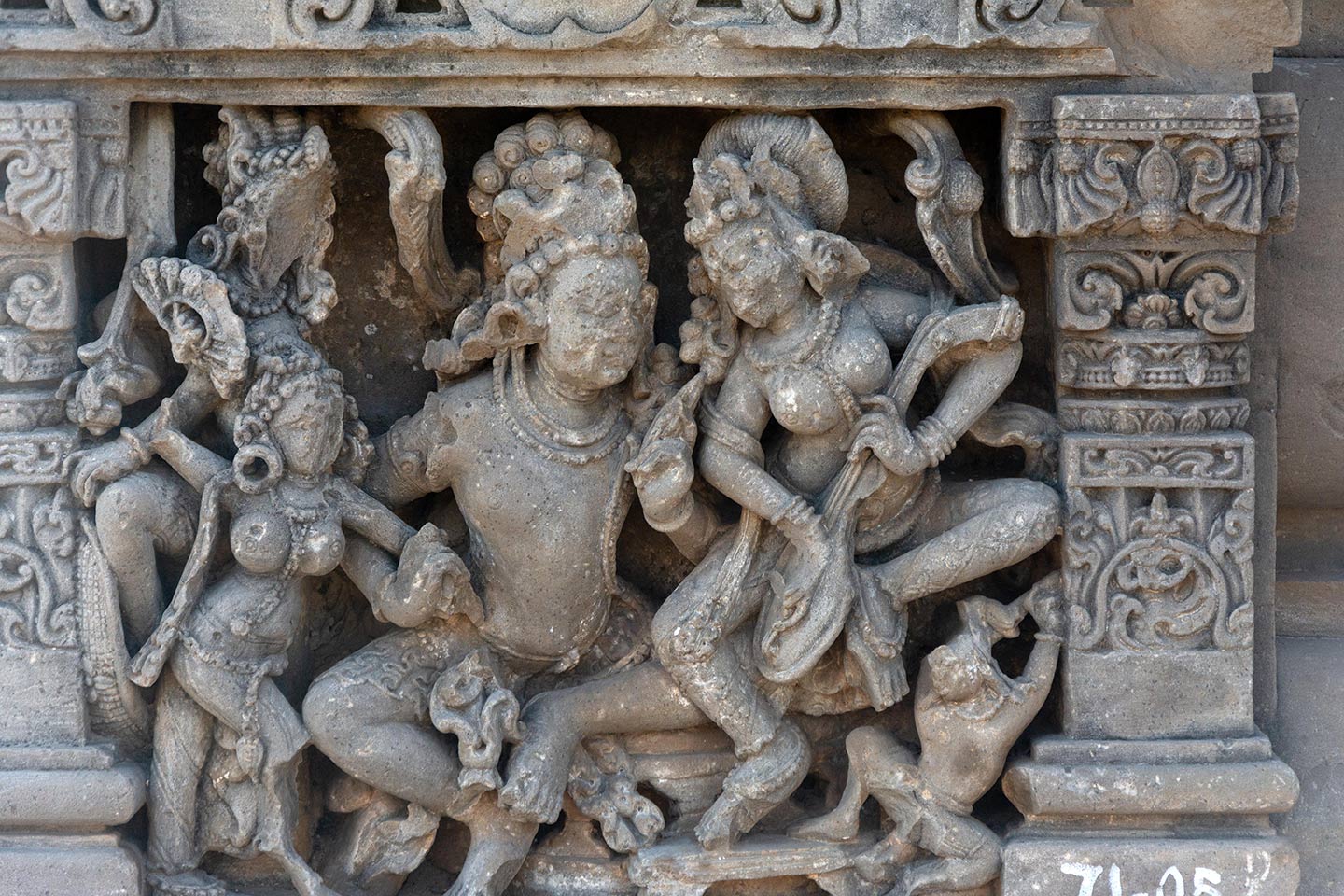 A central male figure is seated in the lalitasana pose on a raised seat, looking towards the central female figure seated on his left thigh, who is playing a stringed musical instrument. The female figure is also in the lalitasana pose and her right foot is resting on a footrest. The central male figure is forcefully pulling a female attendant (holding a hand fan) towards him with his right hand. Behind the fan-holding female is a figure (face missing) wielding a sword.