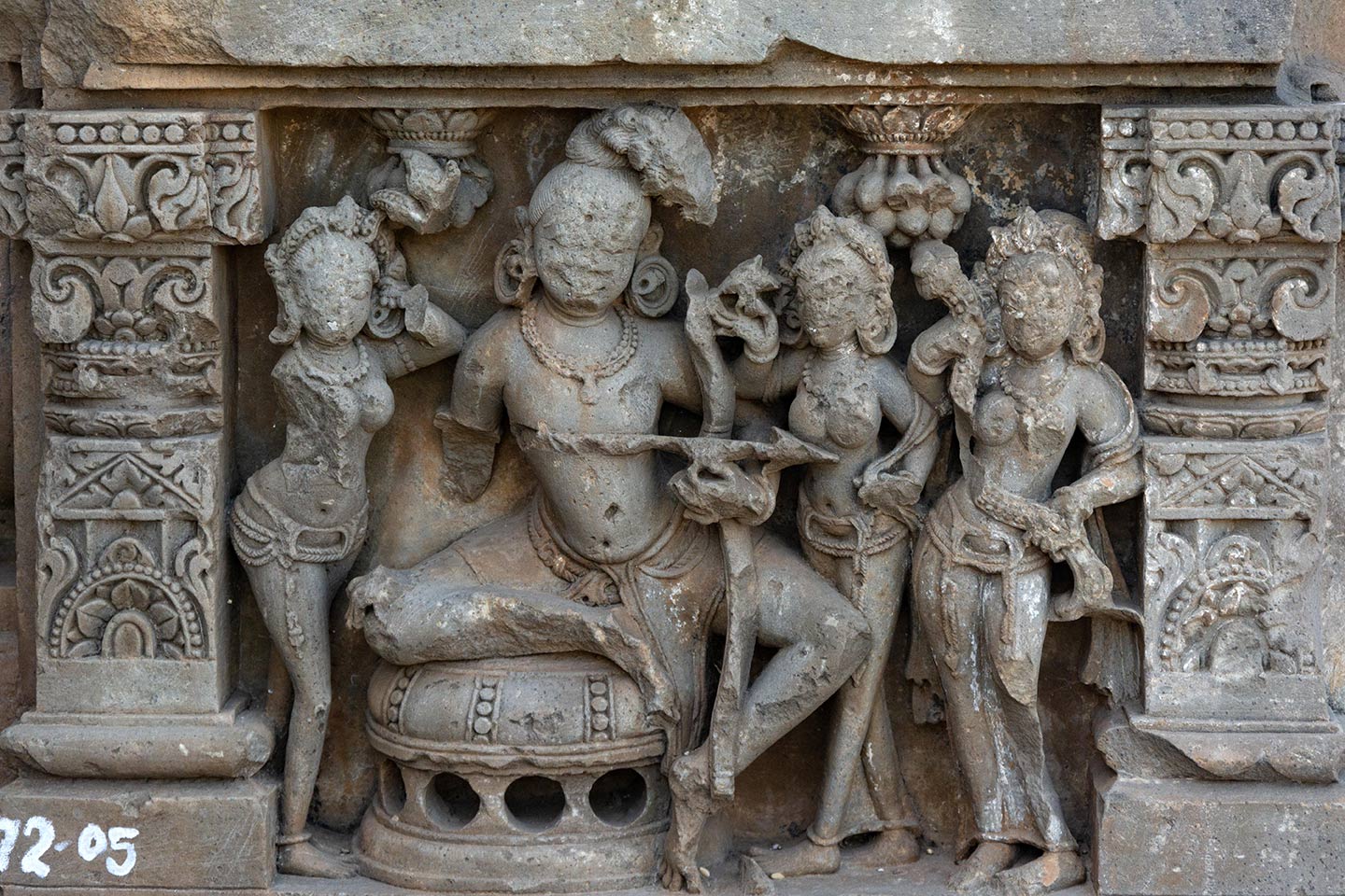 The central male figure is seated on a raised circular seat in the lalitasana pose, firing an arrow from a bow. Standing on his left are two female figures (faces defaced), looking towards him. The female on his immediate left is holding a flower in her right hand. The female to his far left is holding a floral scroll. The female figure on his right is damaged. A bunch of mangoes hang above them.