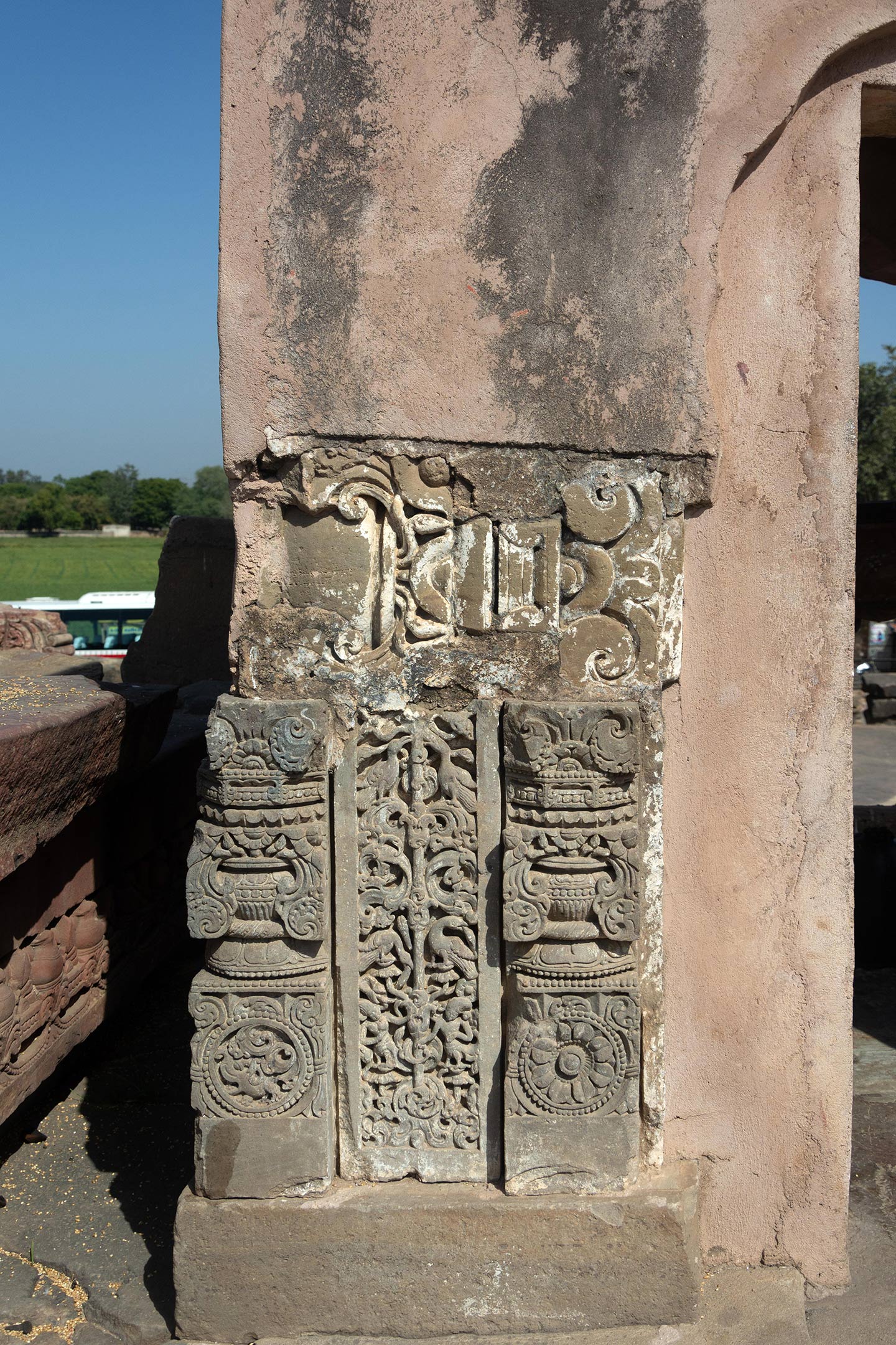 The stone is broken and has three vertical sections. The section on the left has a purna kalasha (overflowing pot of prosperity) motif on the upper half and a makara (hybrid sea creature) inside a circular medallion at the bottom half. The mid-section has an intricate carving of kalpa vrisksha (tree of life) motif with figures of birds and kinnaras (hybrid human-bird creatures) camouflaged within the foliage. The section on the right has a purna kalasha motif on the top half and an eight-petal lotus blooming inside a circular medallion at the bottom half.
