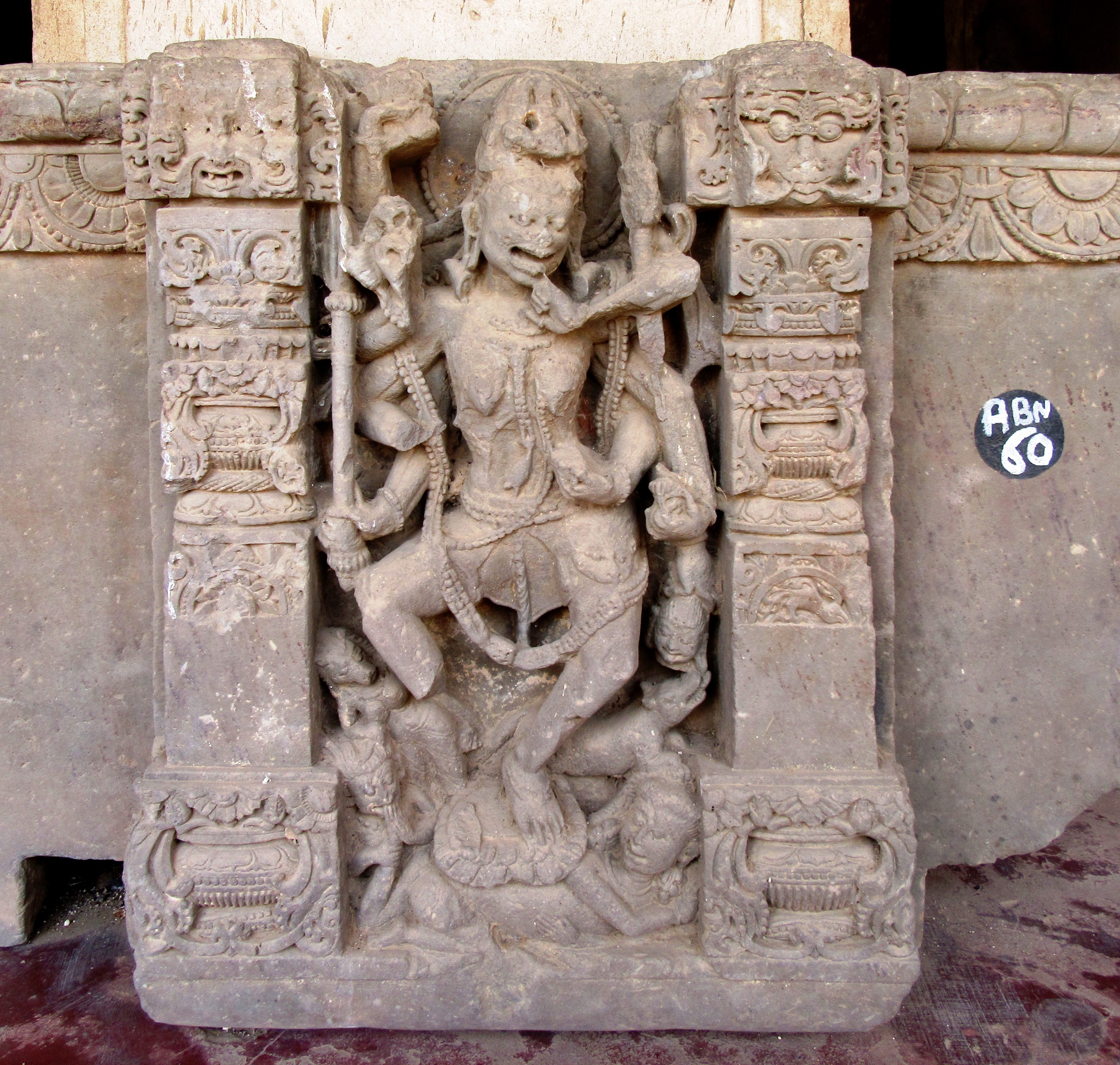 Image 9: An eight-armed goddess stands on a lotus and naravahana (human corpse). Her form appears emaciated with sagging breasts and a sunken stomach. She has her mouth open and is pointing her little finger towards it with her left hand. She holds a trident in her right hand, as well as a khappar (skull bowl) and naramunda (severed head) spear in her left hand. The remaining hands are broken.