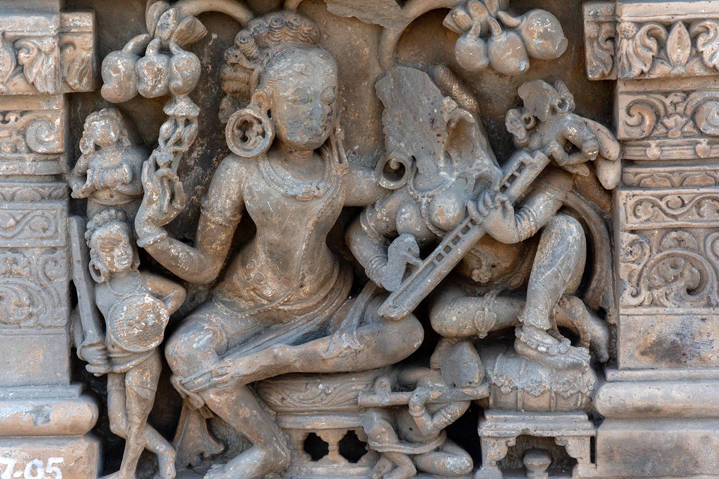 Image 4: The central male figure and female figures are enjoying a musical concert in a grove. Seated on circular raised seats, the male figure assumes a lalitasana pose. The female figure leans towards the male and plays a stringed musical instrument (likely a veena). Four smaller figures surround them. A male soldier is holding a sword and shield (on the left). One musician is sitting between the seats, playing the flute (bottom). A small female attendant is whispering in the central female's ear (top right). Mangoes cling to the tree branches.