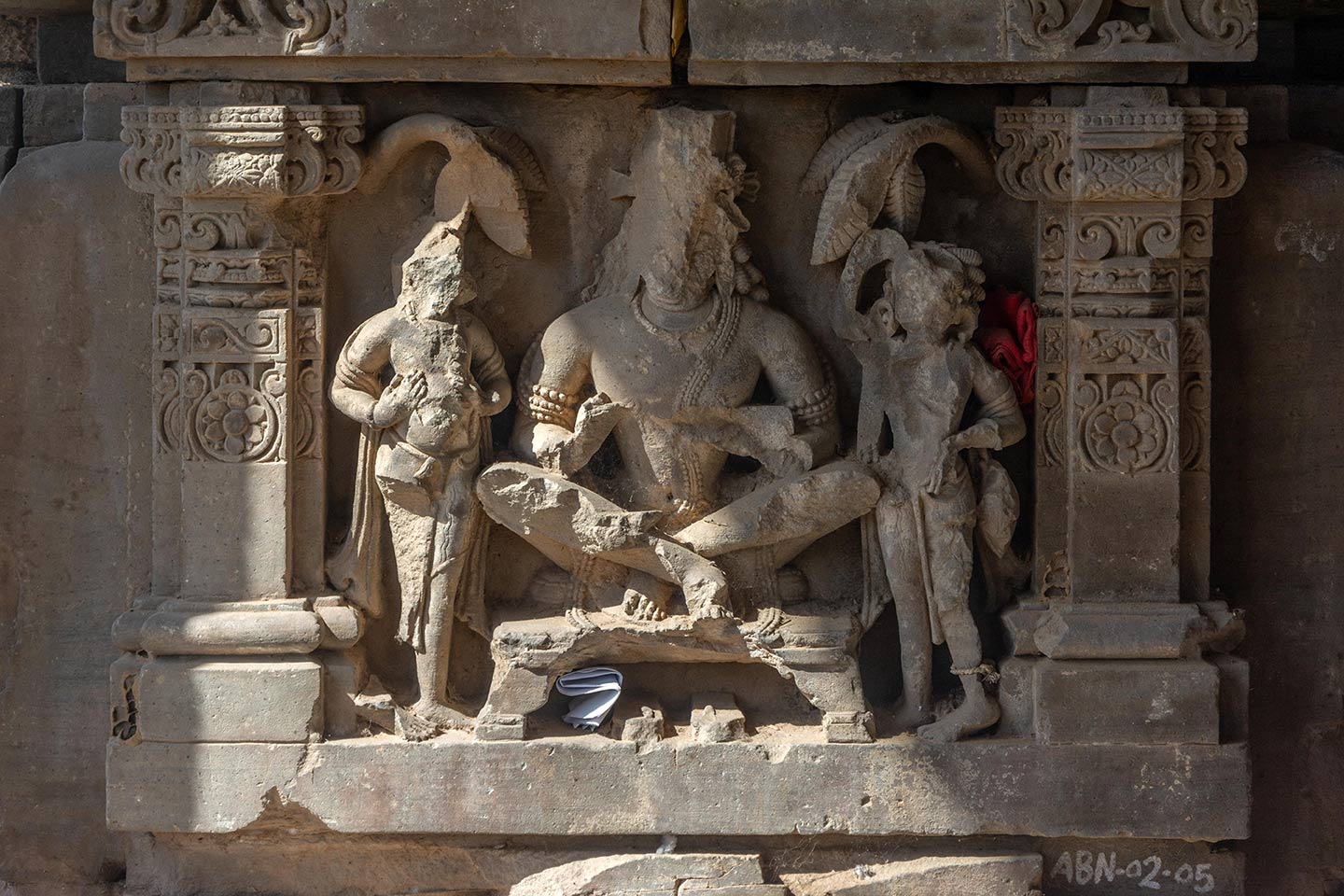 Image 14: This panel depicts a male figure seated in the udichya mudra (cross-legged position). The panel depicts a central male figure seated on a raised asana in the udichya mudra (cross-legged position). A pair of footwear rests at the bottom of his asana. On either side, two female attendants are present, standing in front of trees in the background.