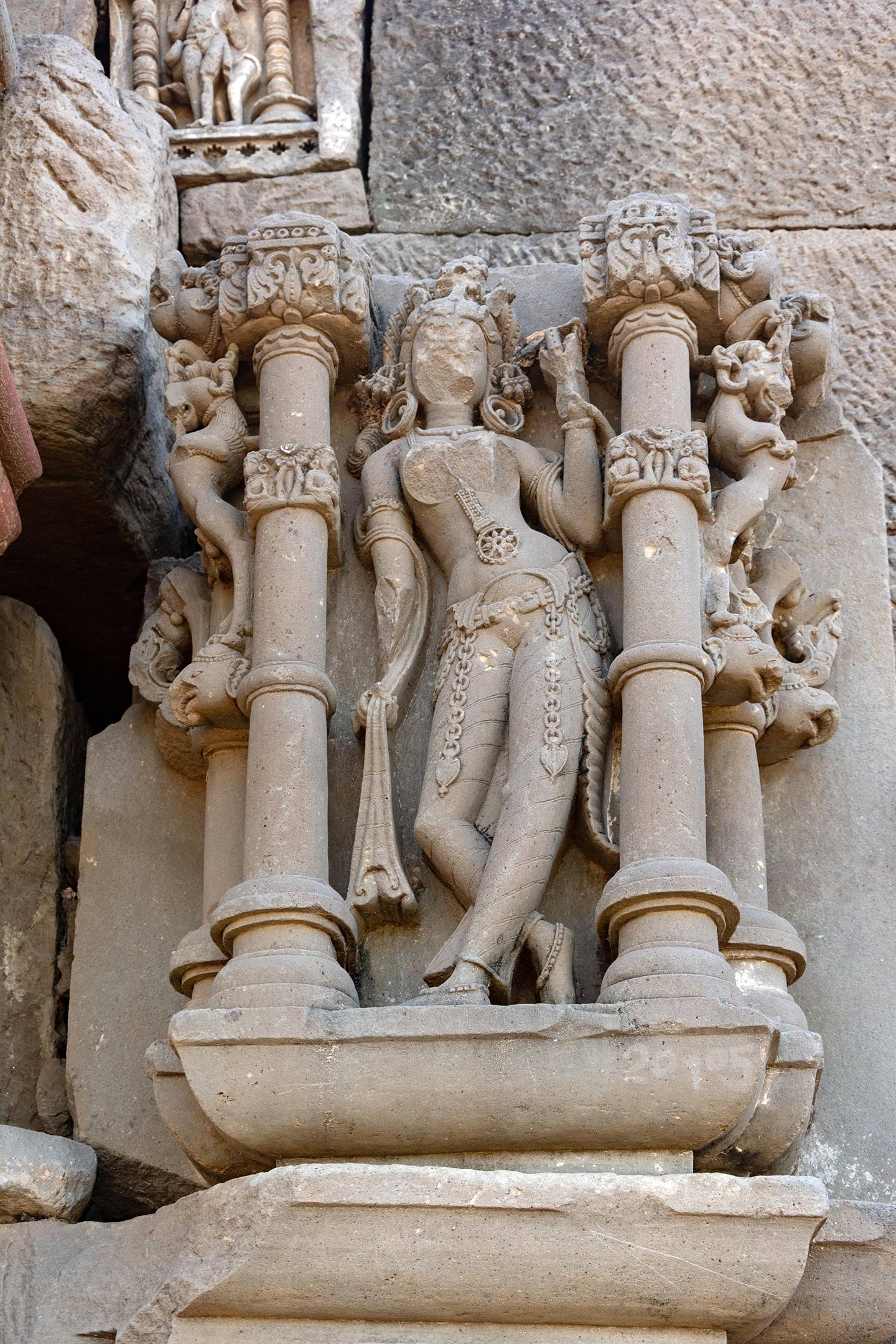 Image 16: The female figure may be an apsara (celestial nymph) or surasundari (young girl as a symbol of graceful beauty) standing in the tribhanga pose (triple-bend). She has a darpana (mirror) in her left hand, and the free end of a dupatta-like garment wrapped around her arms, back, and shoulder in her right hand. Despite the severe damage to her face, the headgear and hoop earrings remain visible. Among the various ornaments she is wearing are a pendant hanging from the navel and two leaf-shaped pendants hanging from chains attached to the katibandha (decorative belt) around her waist.