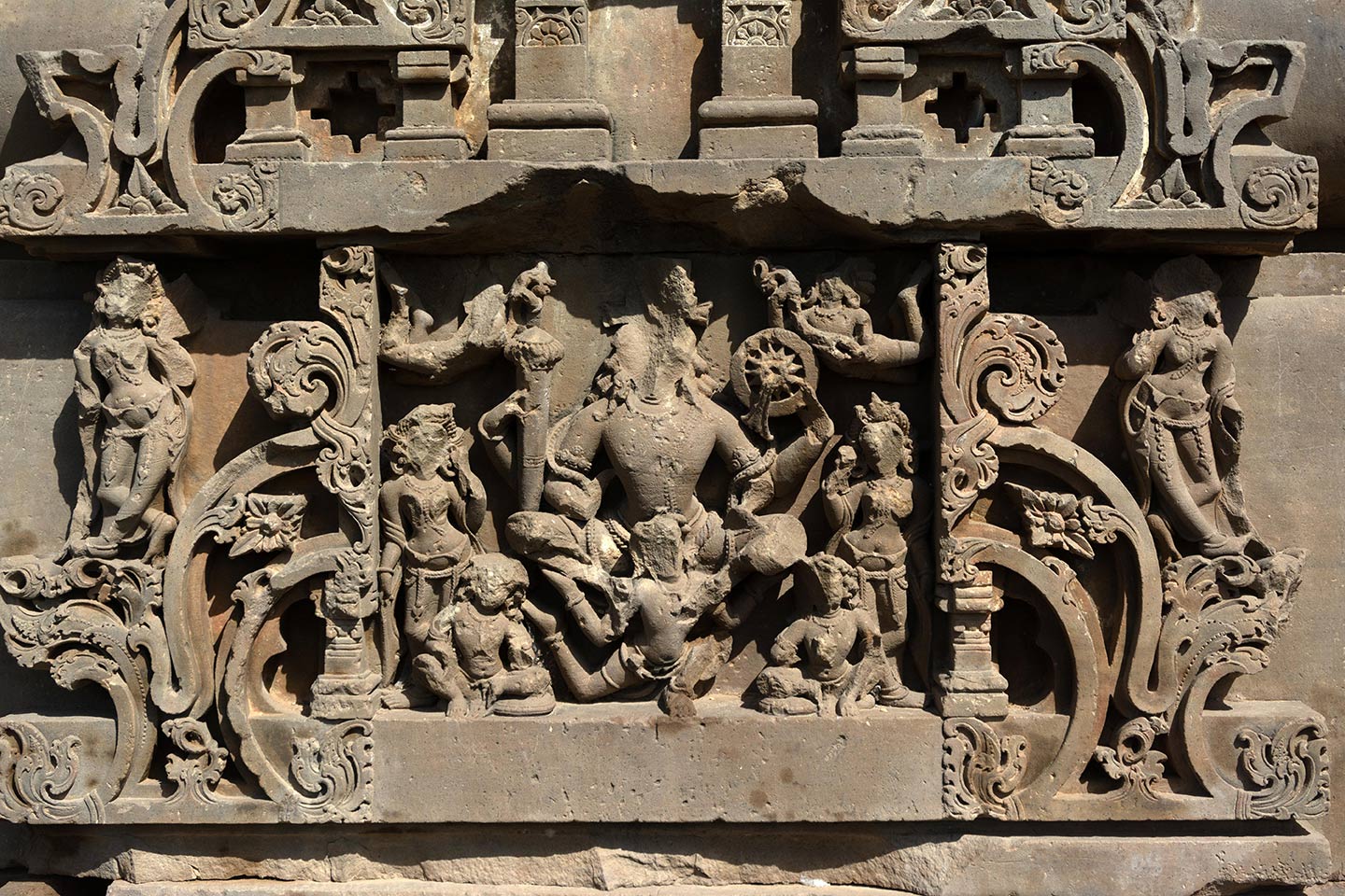 Image 18: This panel depicts Lord Vishnu seated on his mount Garuda. The panel depicts Lord Vishnu as chaturbahu, meaning he has four hands. He carries the gada (mace), named Kaumodaki, in his rear right hand, and the chakra (discus), named Sudarshana, in his rear left hand, while his front two hands remain broken. On the top corners, we see a pair of vidhyadharas (knowledge bearers), showering floral garlands. Two male attendants (seated) and two female attendants (standing) flank Vishnu on either side.